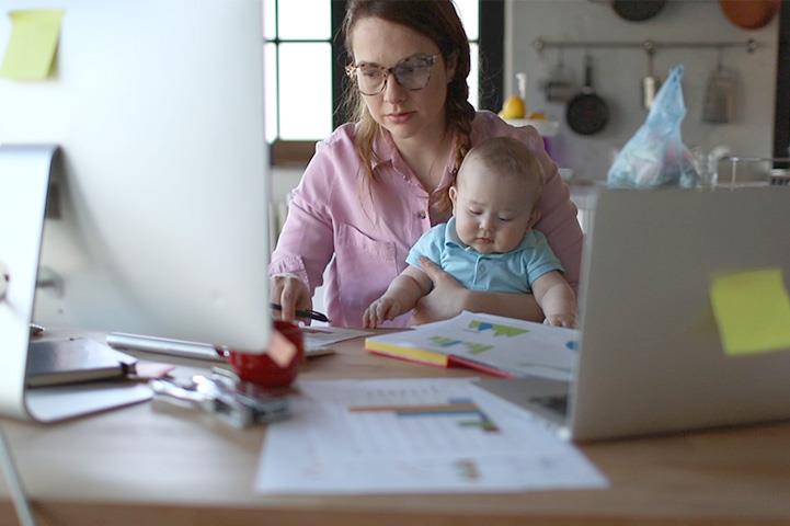 A mother is working at home, on her computer, while holding her baby.