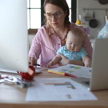 A mother is working at home, on her computer, while holding her baby.