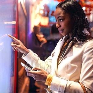 An image of a woman standing in front of a touchscreen holding a phone.