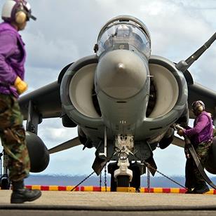 one man in bdu walking in front of a harrier jet while another man adds fuel