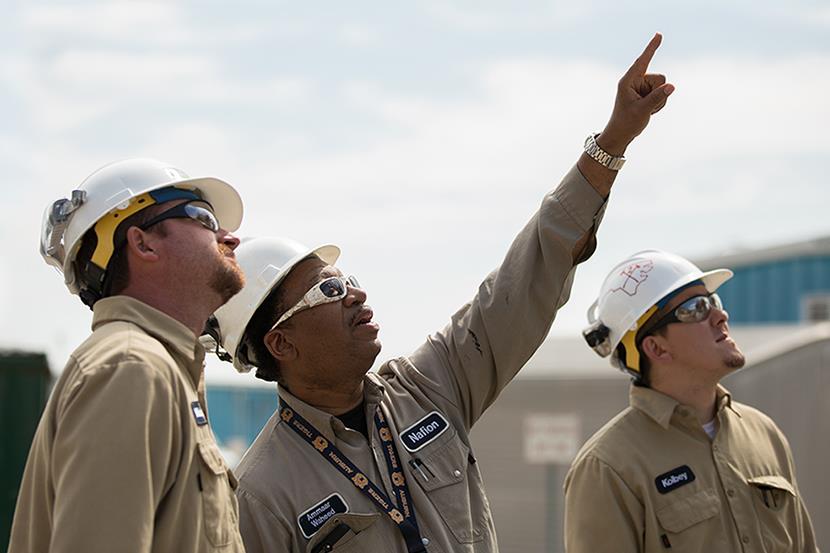 three men in white hardhats tan coveralls and sun glasses looking up at something the middle man is