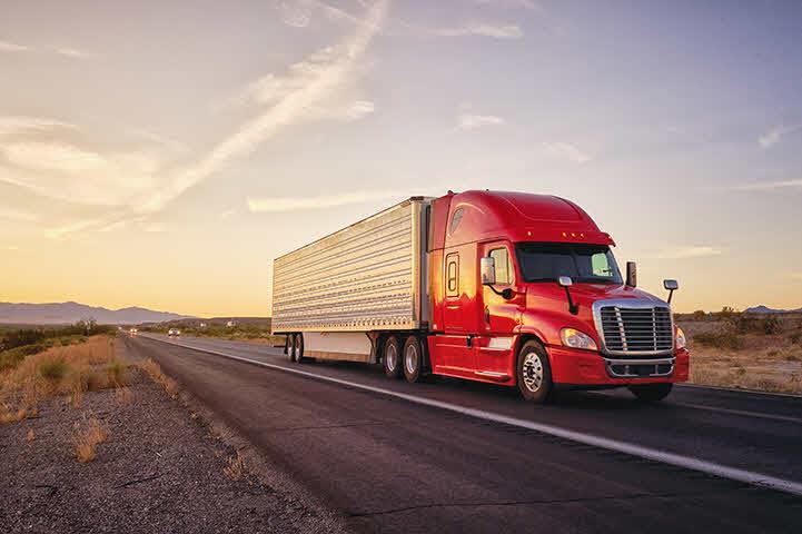 An image of a red tractor trailer driving down the highway near sunset.