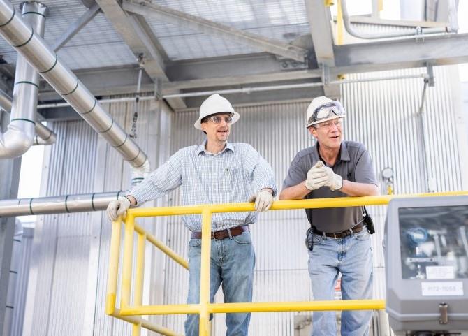 two men wearing hardhats leaning against a railing in a factory